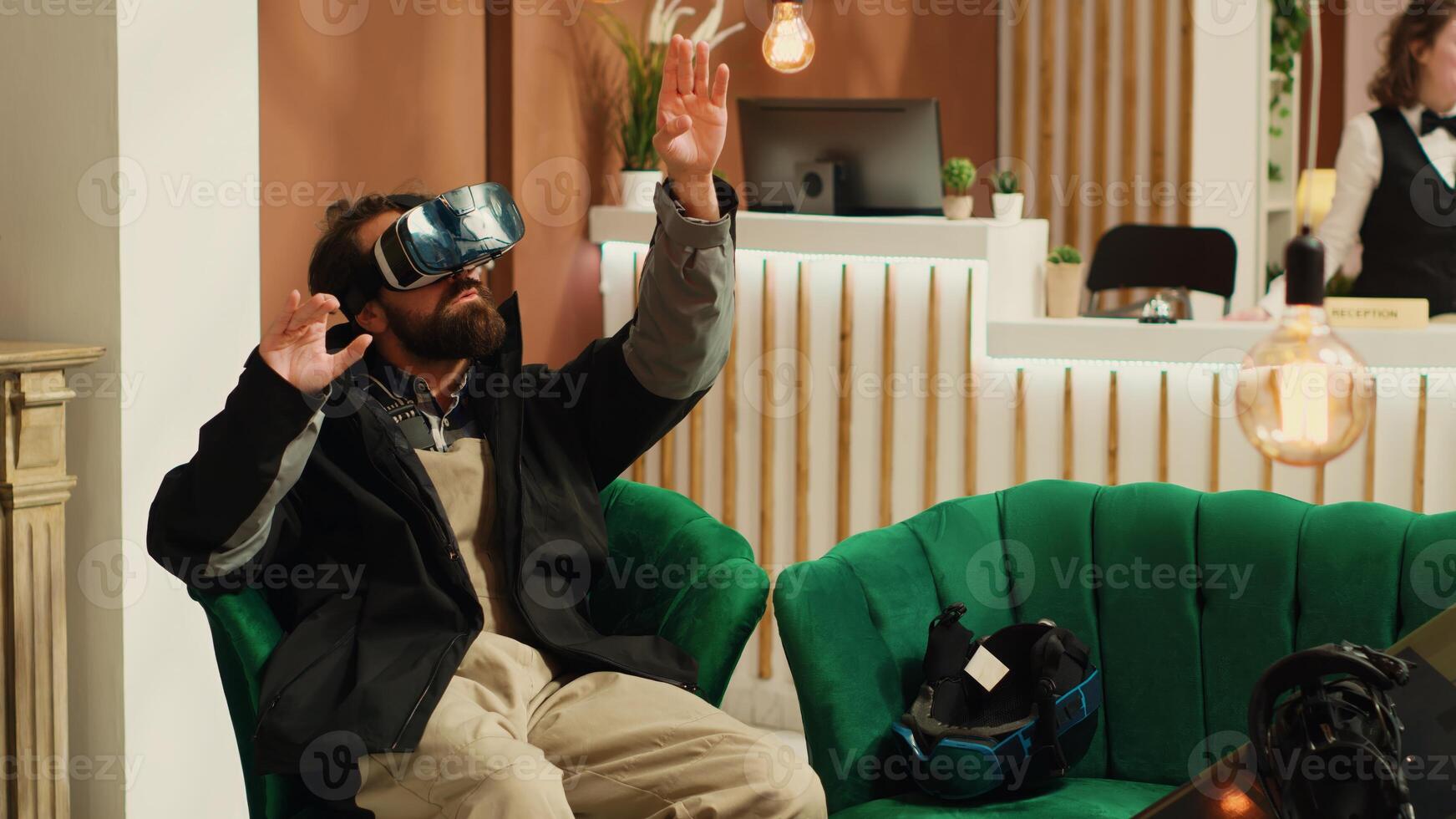 Traveler uses virtual reality technology in lounge area, having fun with vr interactive glasses while he wears winter jacket. Person having helmet and snowboard next to him, prepare to go skiing. photo