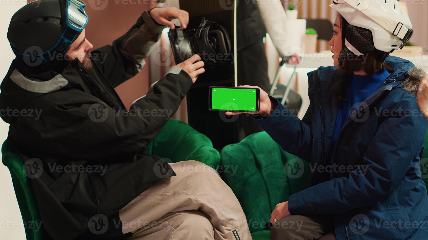 Woman holds smartphone with greenscreen while she talks to friend about winter sports at mountain ski resort. People equipped with skiing gear and protection, isolated mockup template. photo