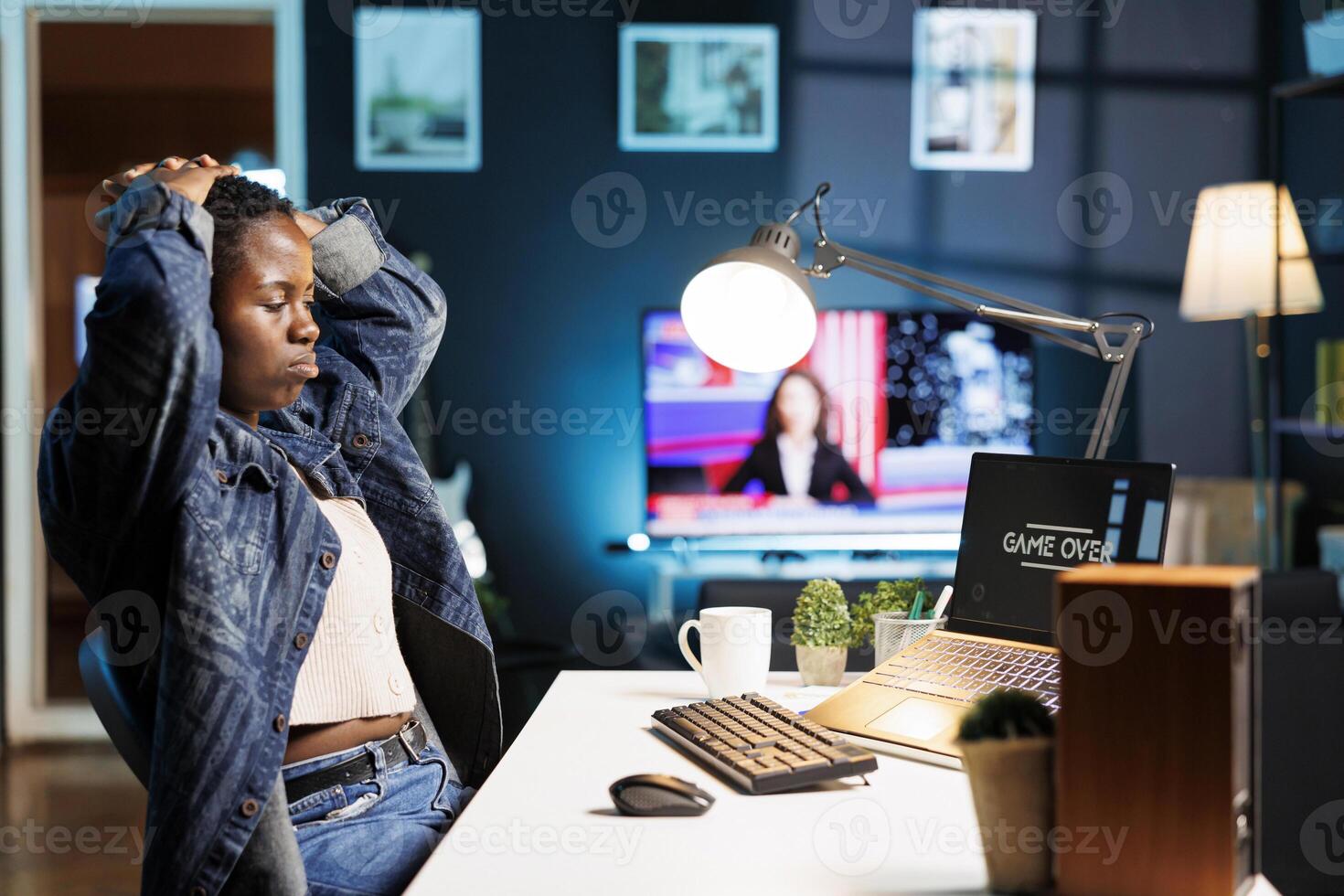 Black woman with hands on head sadly looking at laptop screen showing game over. Upset african american female gamer sitting at desk in apartment after losing video game on portable computer. photo