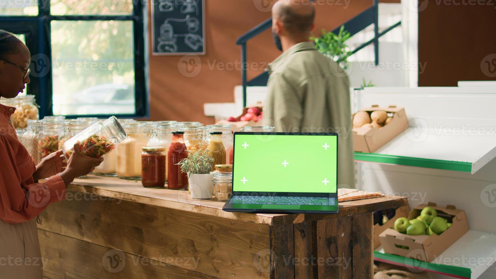 Greenscreen on device in eco store with crates of fresh homegrown products and bulk items in jars on shelves. Laptop showing isolated template with mockup chromakey display. photo