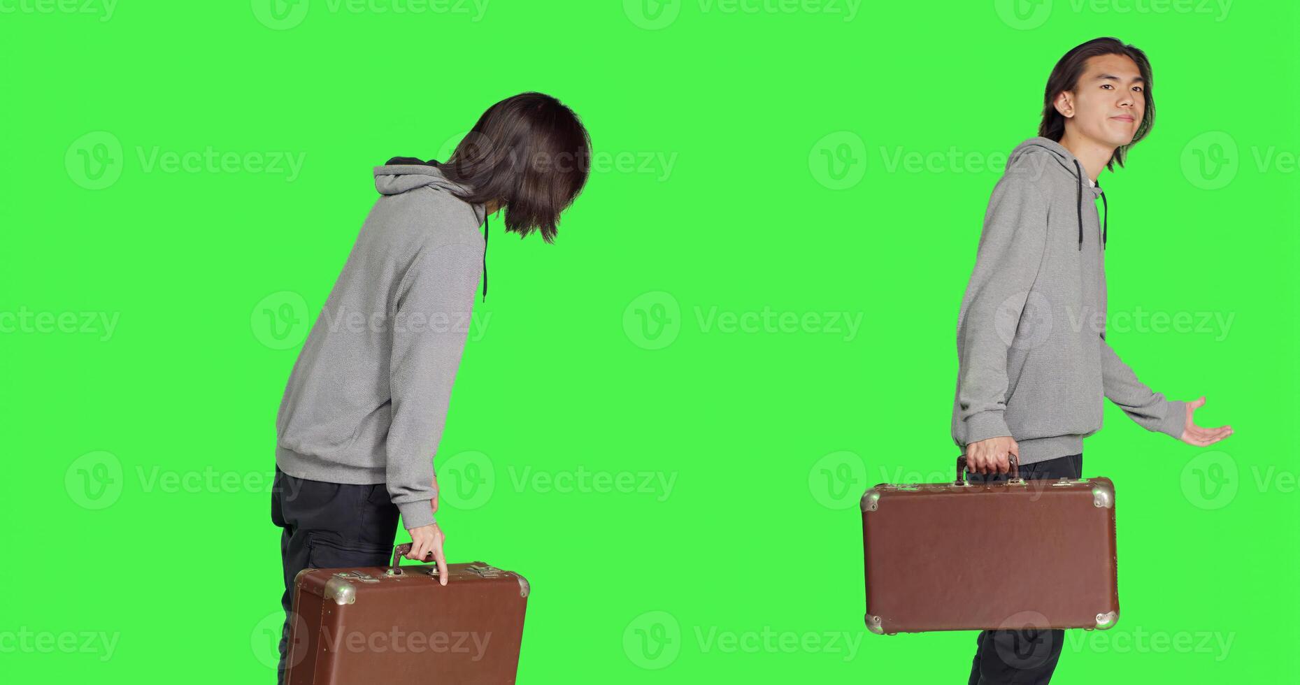 Young adult holding vintage suitcase over greenscreen backdrop, waiting impatiently for a certain thing. Asian man carrying huge briefcase while he looks at time on wrist watch in studio. photo
