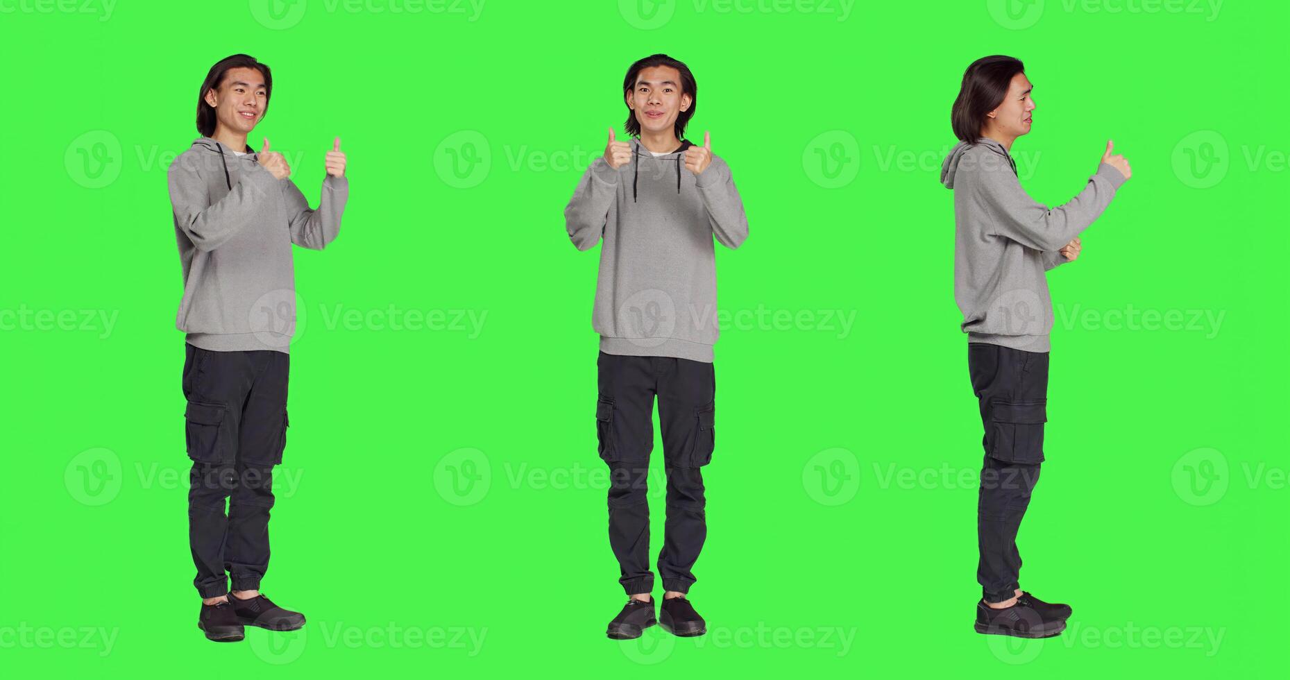 Asian guy does thumbs up gesture standing over full body greenscreen, showing agreement and presenting like symbol in studio. Young joyful adult feeling pleased, approval sign. photo