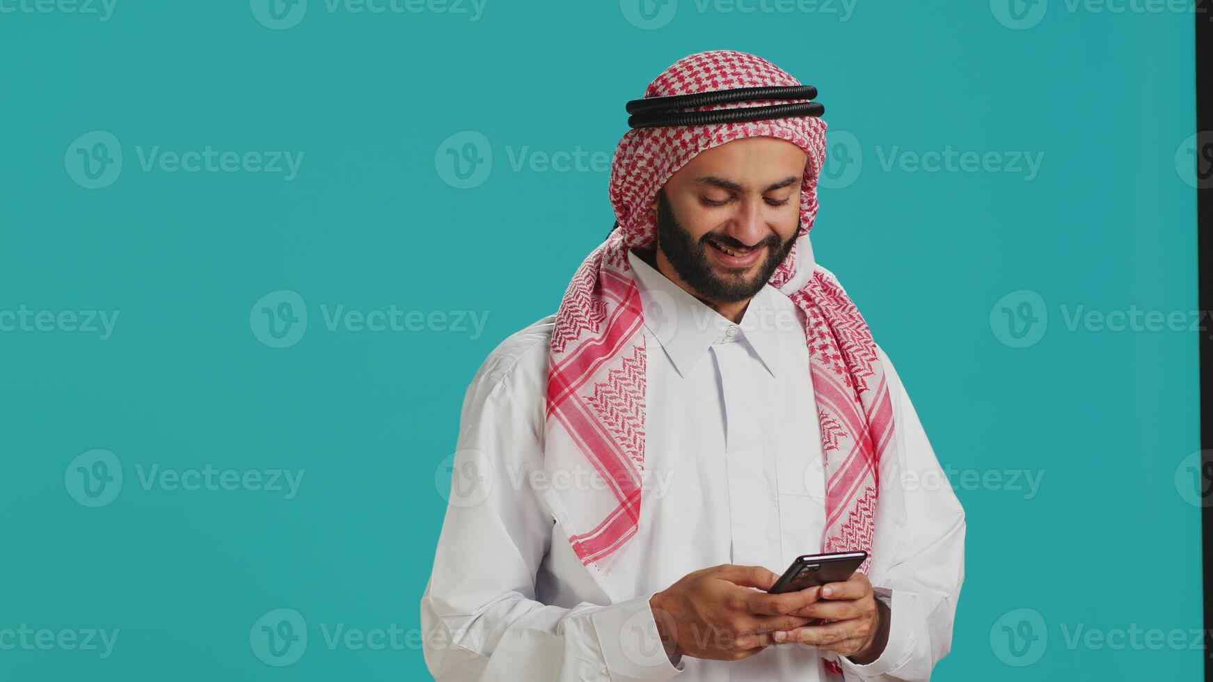 Smiling adult typing messages on phone, wearing muslim traditional clothing. Islamic arab person dressed in white thobe and checkered ghutra chatting on social media webpage, texting. photo