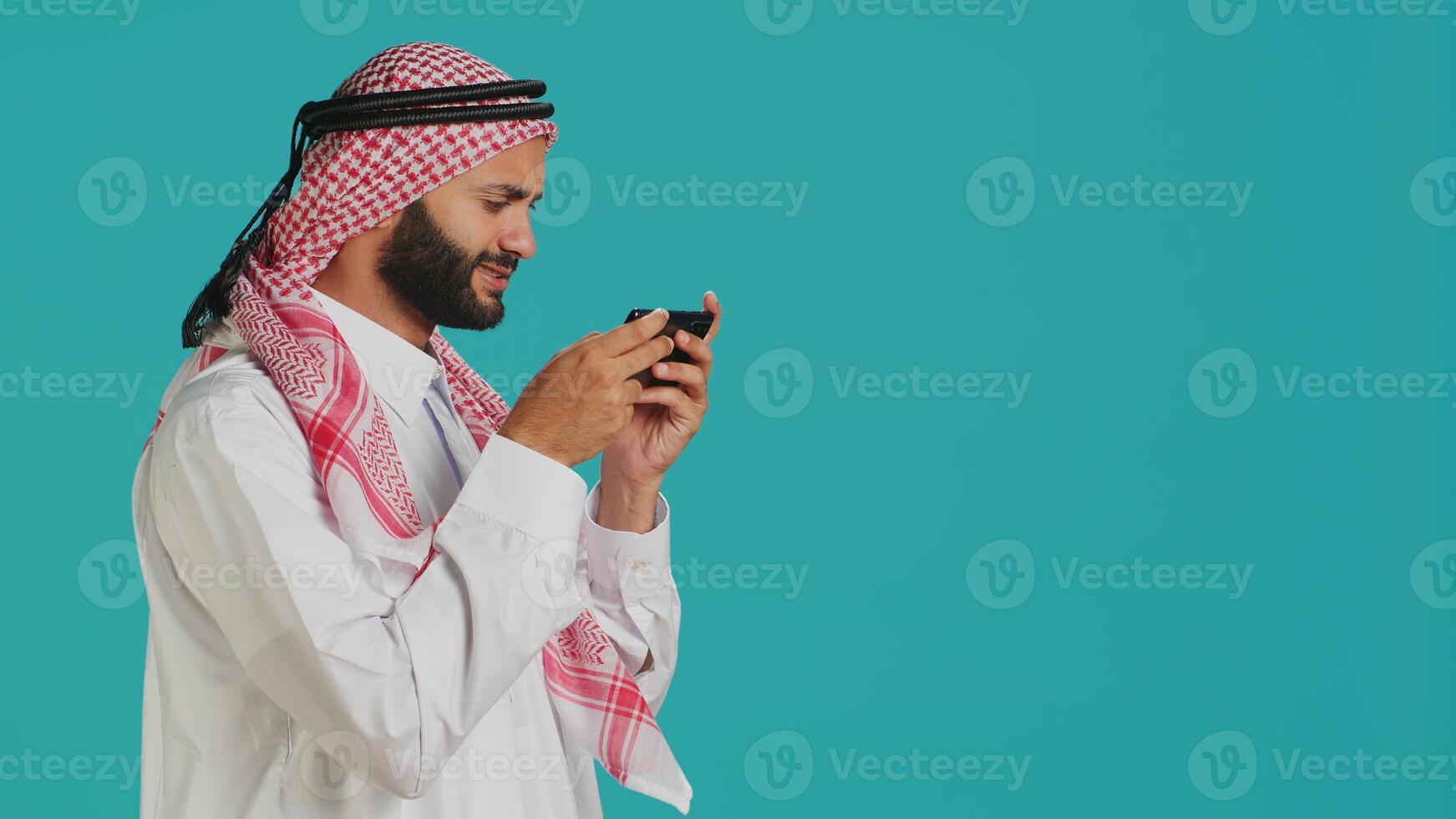 Arab man playing fun mobile videogames on phone, dressed in traditional attire with dress and headscarf, leisure activity. Middle eastern guy engaging in online gaming contest using device. photo