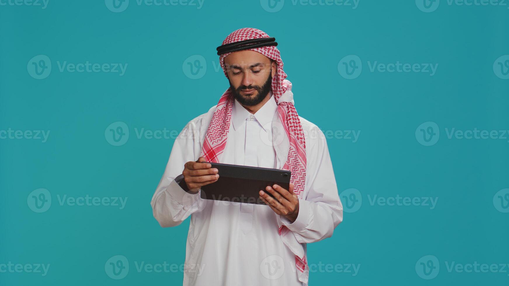 Middle eastern man checking tablet in studio, browsing websites online on digital gadget. Male person in arabic clothes using device to navigate on internet pages, texting on social media. photo
