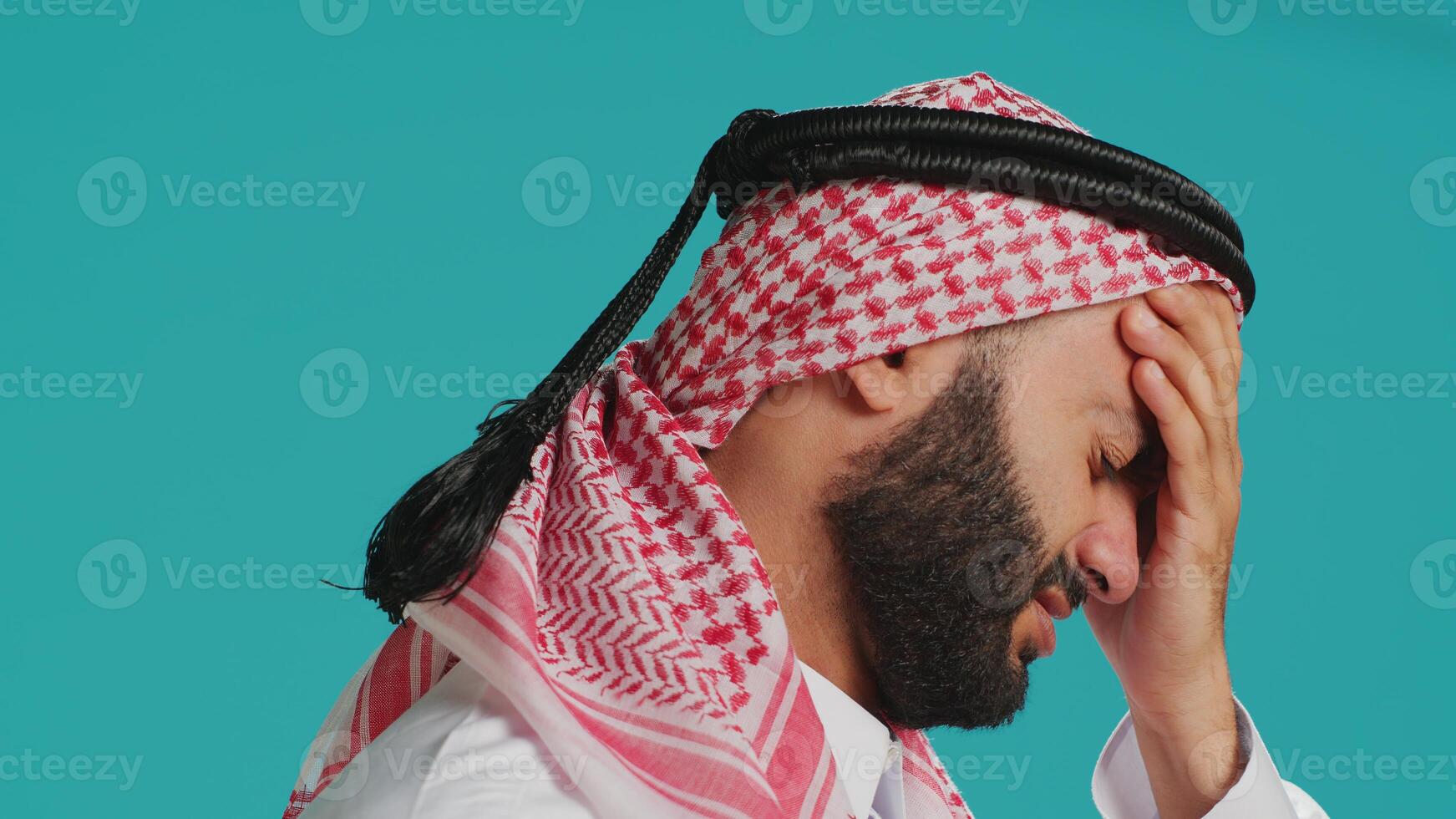 Middle eastern man experiencing awful headache and stress, suffers with migraine over isolated backdrop. Person wearing traditional muslim apparel, feeling ill and worried with sinus discomfort. photo