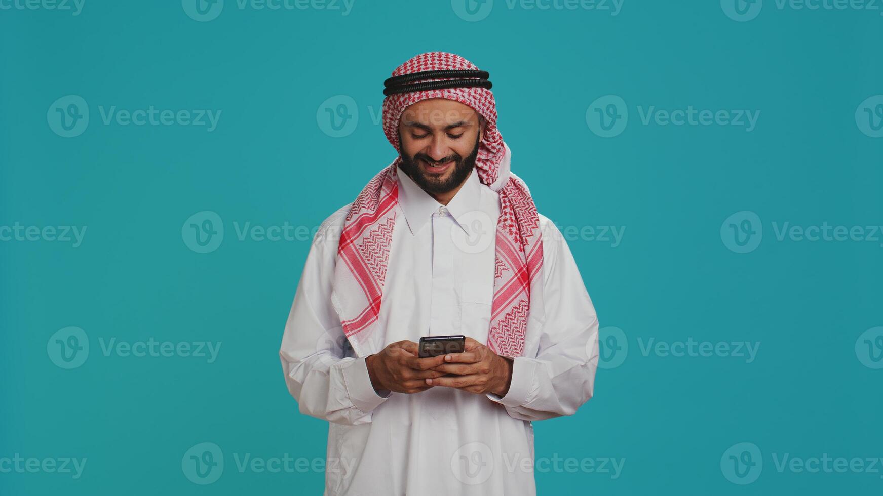 Cheerful person messaging on phone, wearing islamic religious attire and headscarf. Muslim guy dressed in traditional thobe and checkered print ghutra chatting on social media network. photo