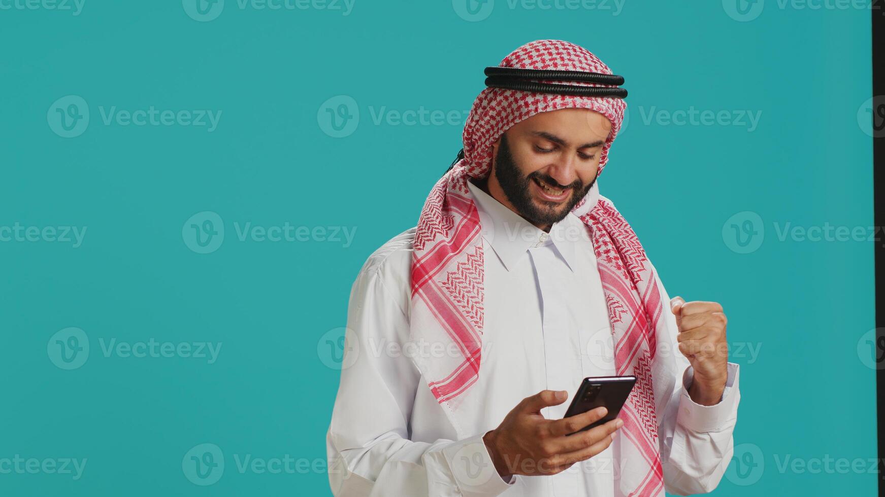 Arabic person in traditional outfit holding smartphone in hands and typing in online social media chat. Muslim adult standing with mobile phone and texting on webpage, internet site. photo