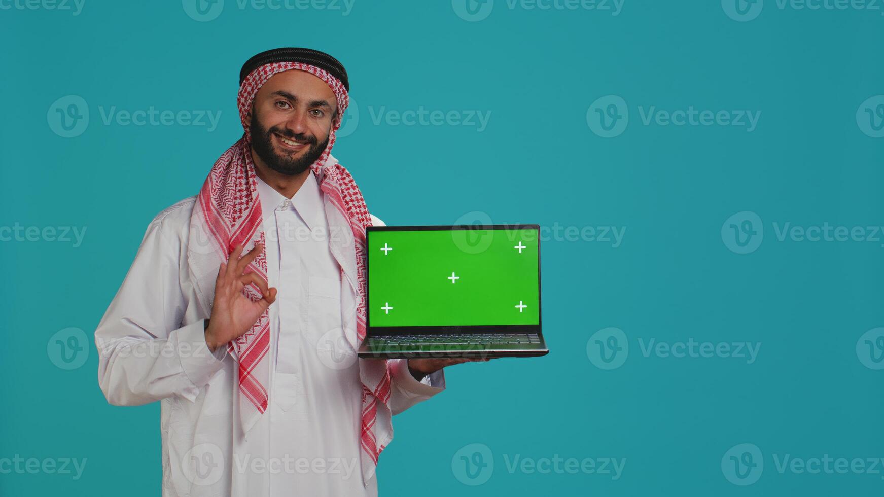 Muslim adult shows greenscreen laptop with blank mockup layout on display, copyspace template. Arab guy wearing gown and headscarf, presenting isolated chromakey screen on pc. photo