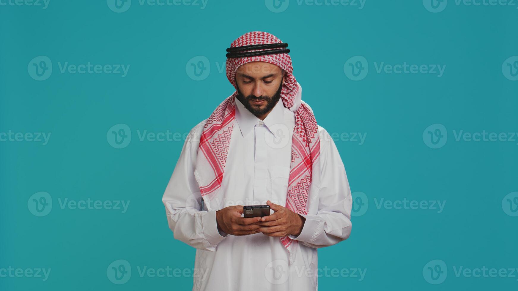 Muslim man browsing smartphone app and feeling cheerful after finding out about great news, typing texts on social media website. Middle eastern guy scrolling online pages, traditional clothes. photo