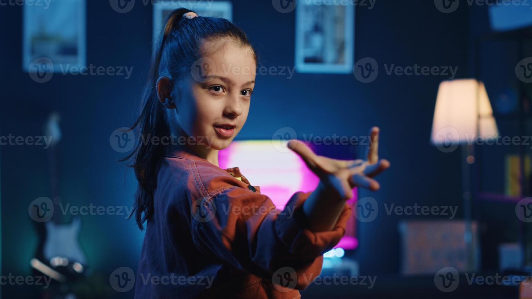 Close up shot of smiling kid dancing in dimly lit home studio interior, producing content for online channel. Excited child doing viral dance choreography in apartment illuminated by neon lights photo