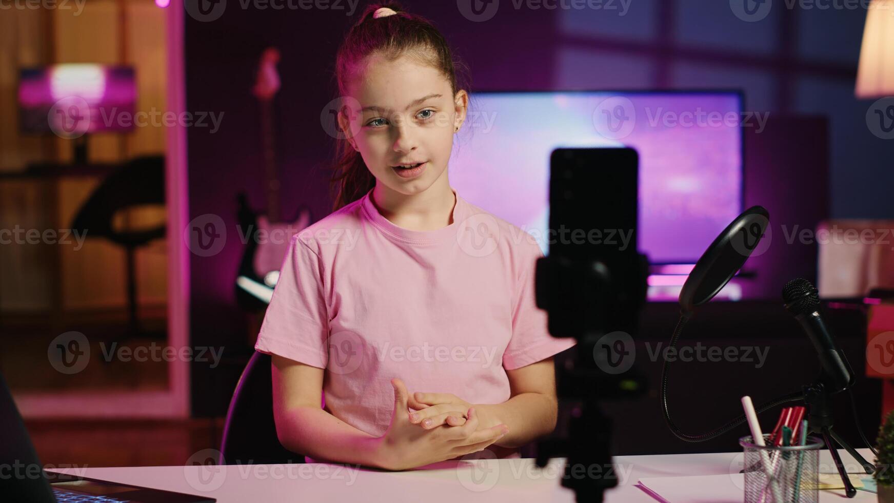 Kid filming with smartphone on tripod stabilizer in pink neon lit living room used as professional studio. Cheerful girl captures footage with mobile phone camera, discussing about fun day at school photo