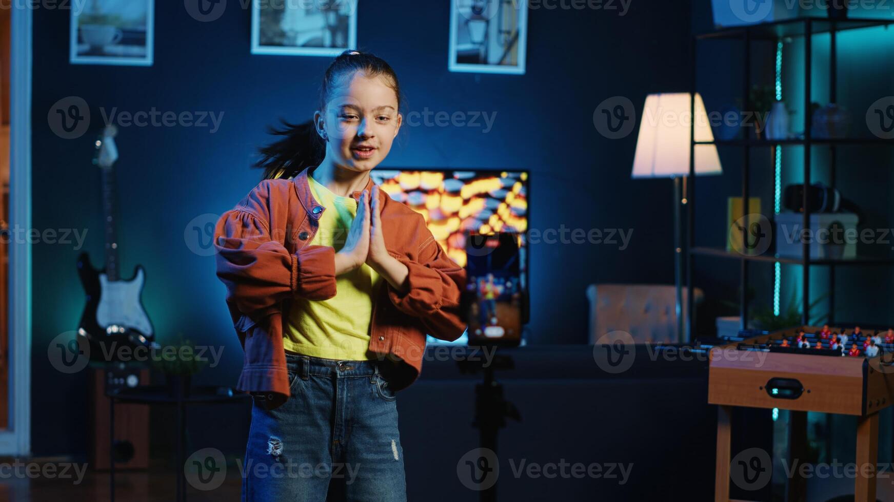 Little girl doing nice dance moves, filming viral challenge using smartphone. Happy kid having fun recording video with mobile phone for online fans in dimly lit home studio photo
