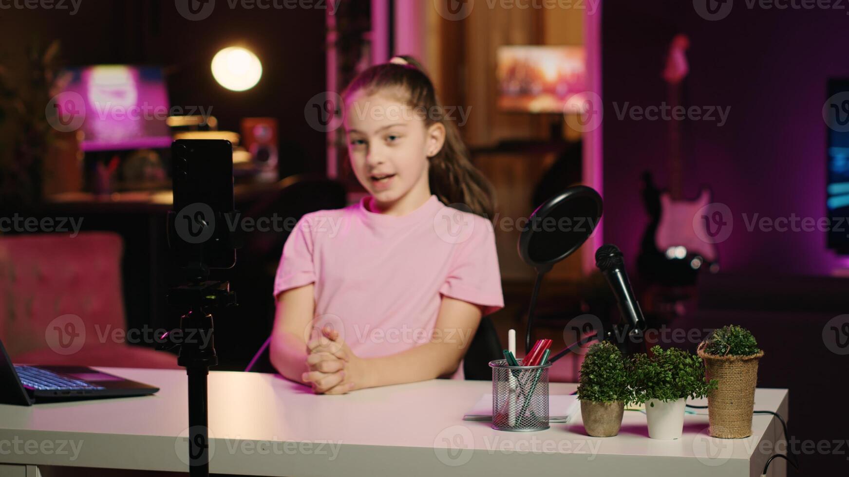 Young girl filming content for her online fans, talking about gen Z topics. Cute child recording vlog for family channel in apartment studio with pink neon lighting as background photo