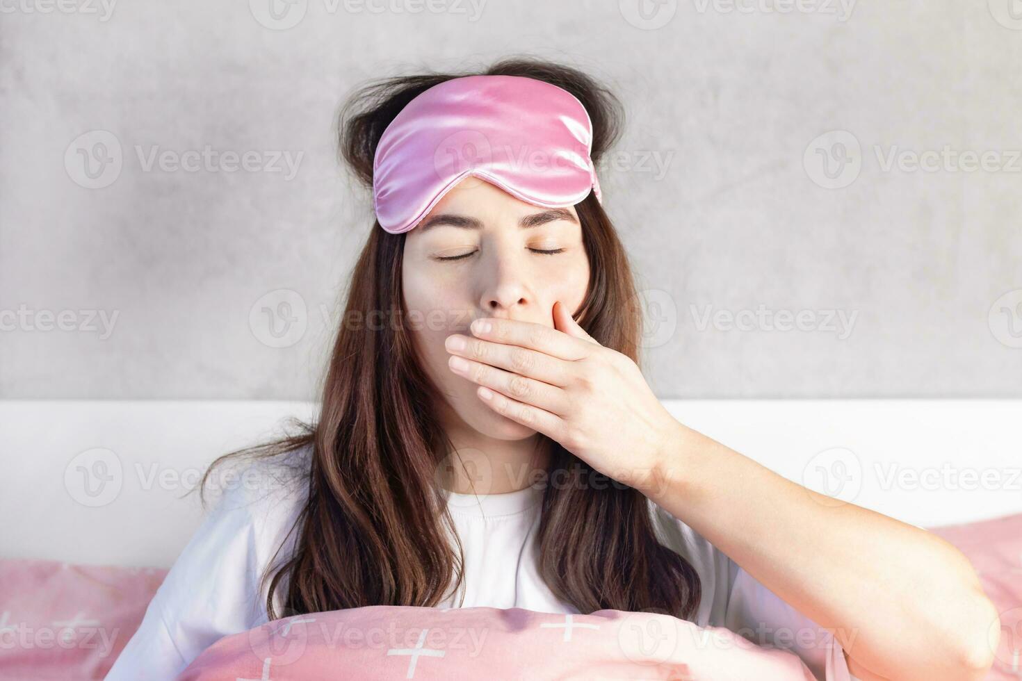A beautiful girl with blue eyes sits in bed in pajamas and a sleep mask. Woman yawns in bed. She wants to sleep photo