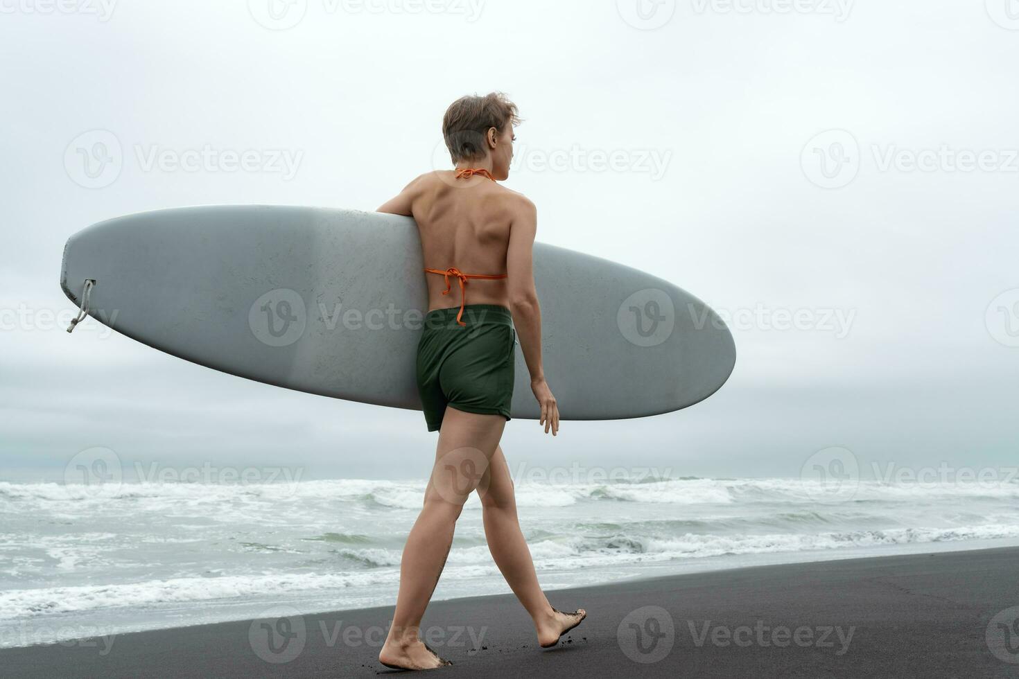 Rear view of female surfer walking on sandy beach carrying surfboard against background of ocean photo