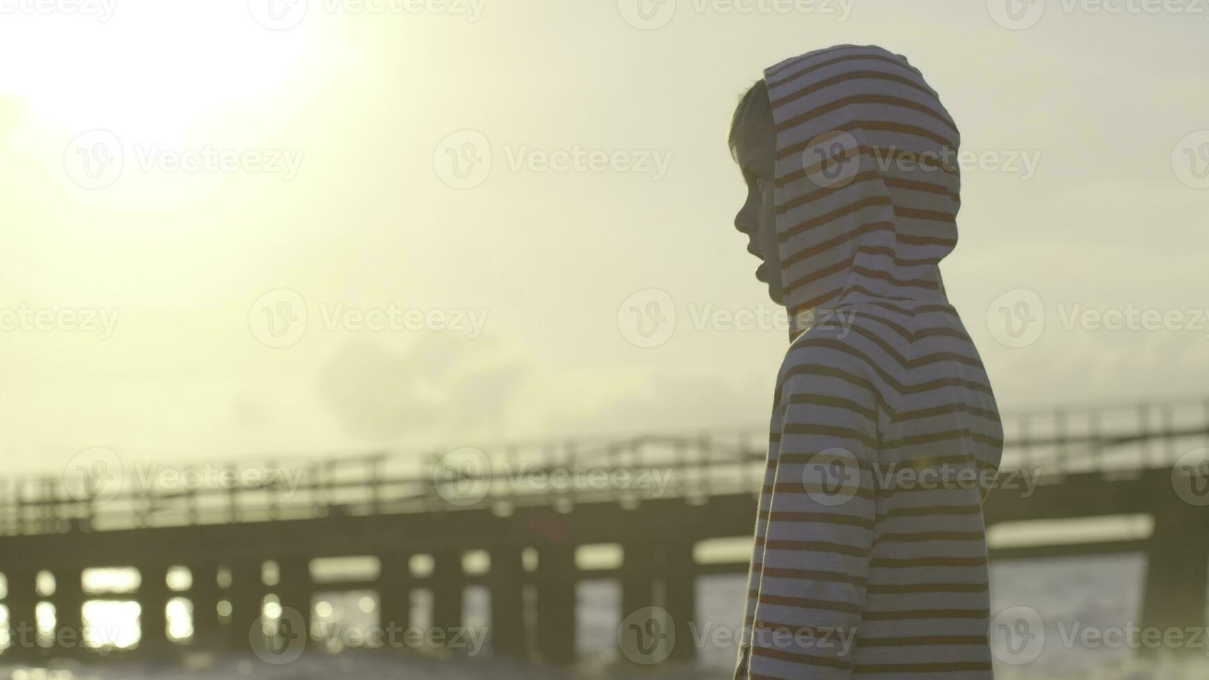 Boy looks at sea on background of pier. Creative. Boy looks at horizon during sunset on beach. Child looks in amazement at sea at sunset in summer photo