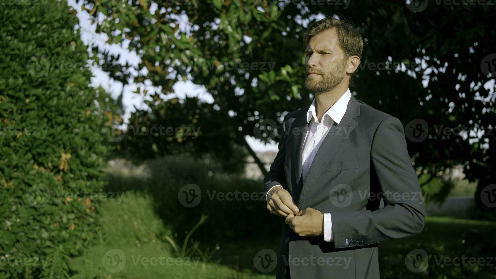 Portrait of young groom posing before wedding ceremony outdoors. Action. Thoughtful man in black and white suit among bushes and trees. photo