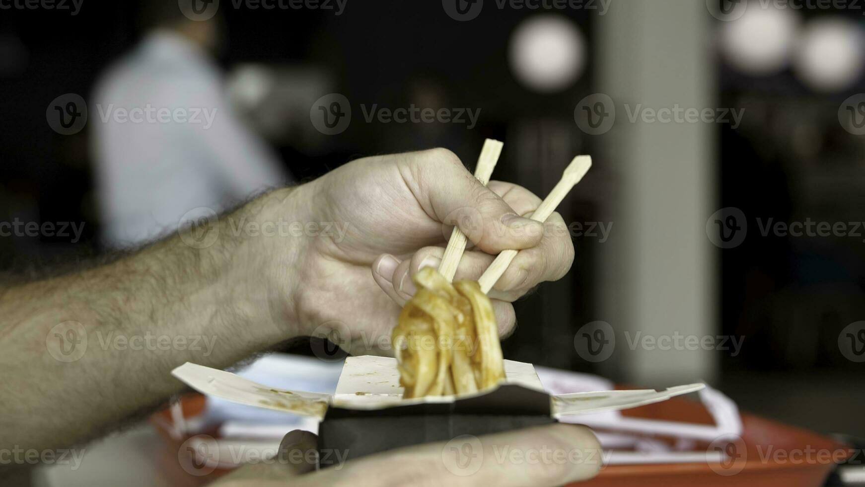 Close up of male hands with wooden chinese chopsticks eating delicious asian wok noodles. Media. Side view of eating chinese pasta in a public place. photo