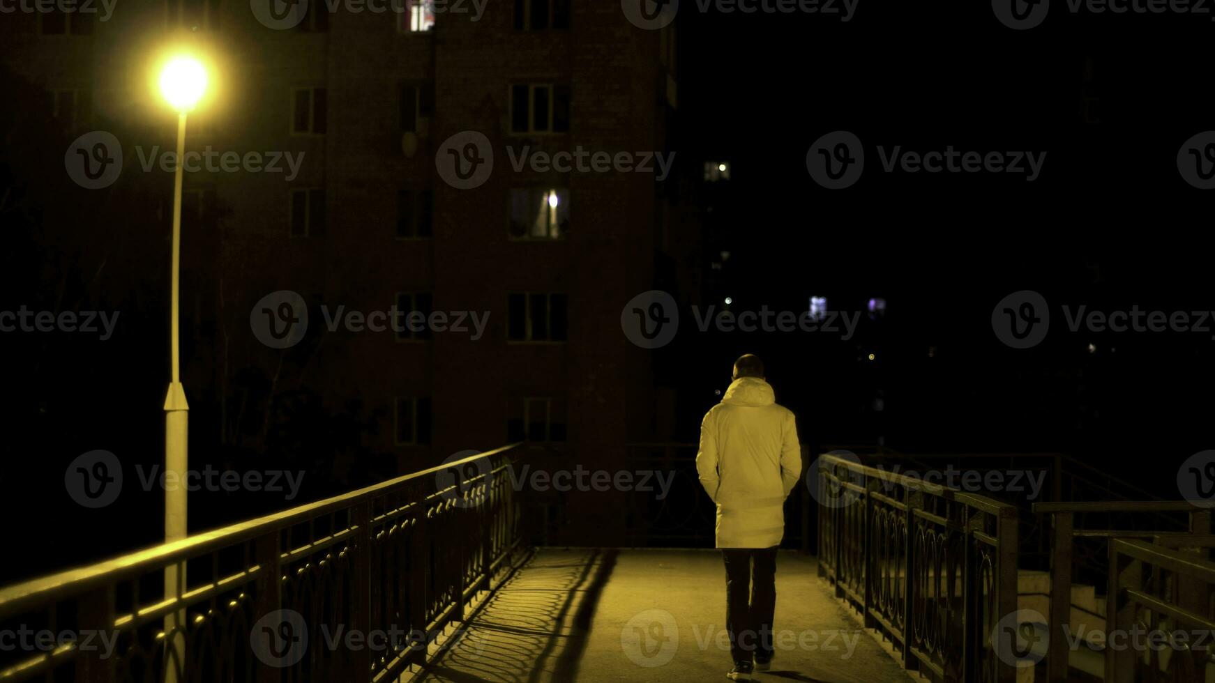 Pedestrian bridge with a lonely street lamp and a man crossing it at night. Stock footage. Rear view of a young man in white jacket crossing the bridge in the city at night. photo