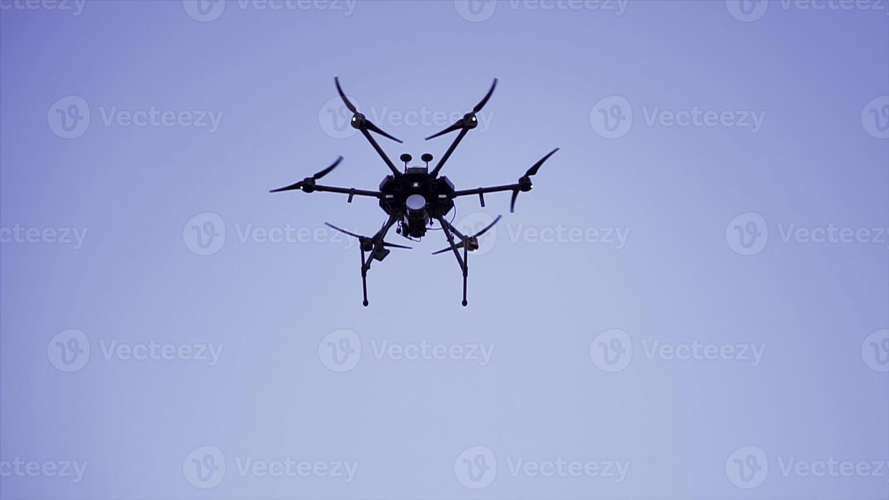 Quadcopter drone hovering in a clear blue sky, video and photo shooting concept. Clip. Black small quadrotor with digital camera and rotating blades, bottom view.