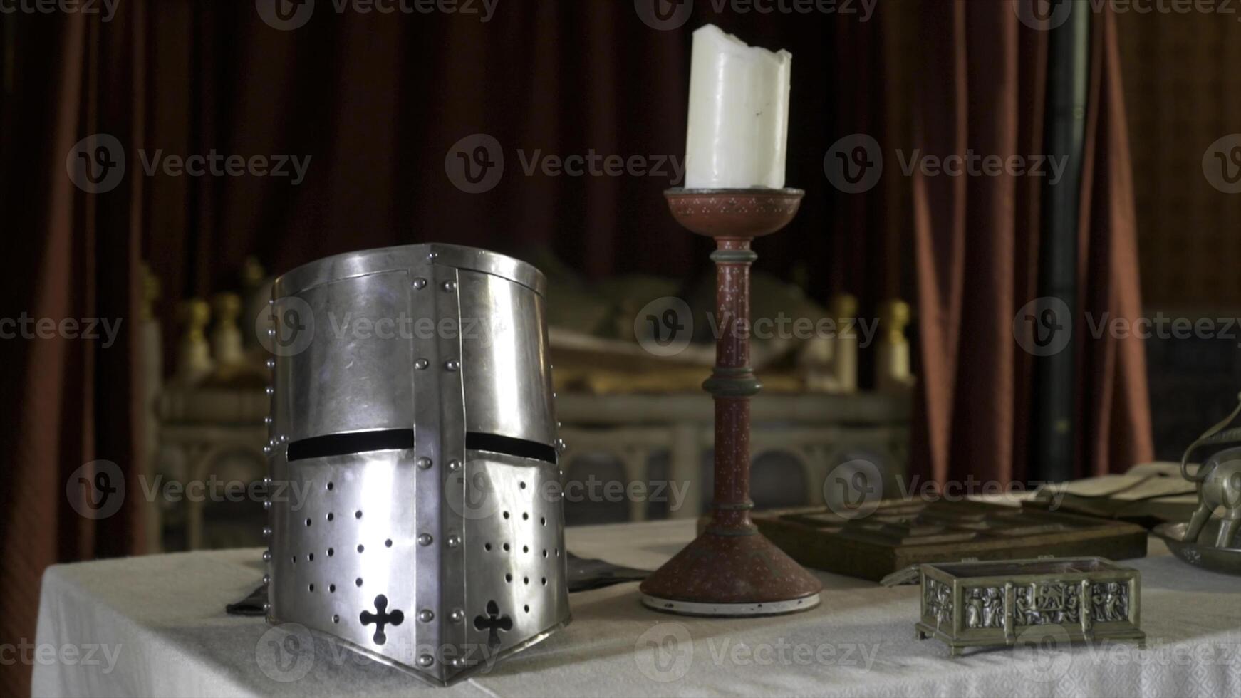 Medieval interior with details and objects. Action. Silver knight's helmet and candle stand on table in medieval interior. Objects of middle ages are on table on background of red fabric in Museum photo