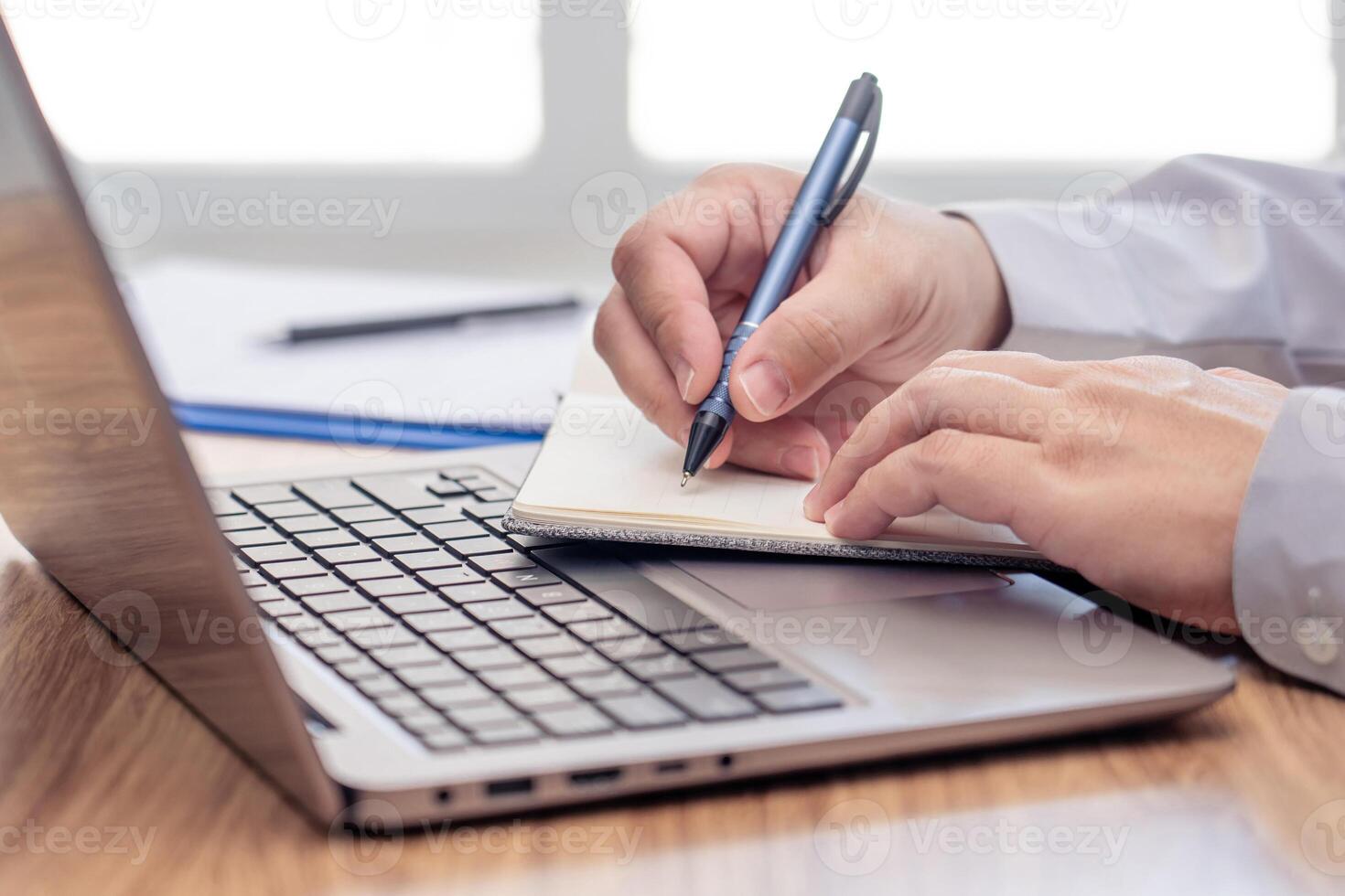Business man write on notebook paper and use laptop computer working and analyzing with document data document for new project of business strategy at workplace. photo