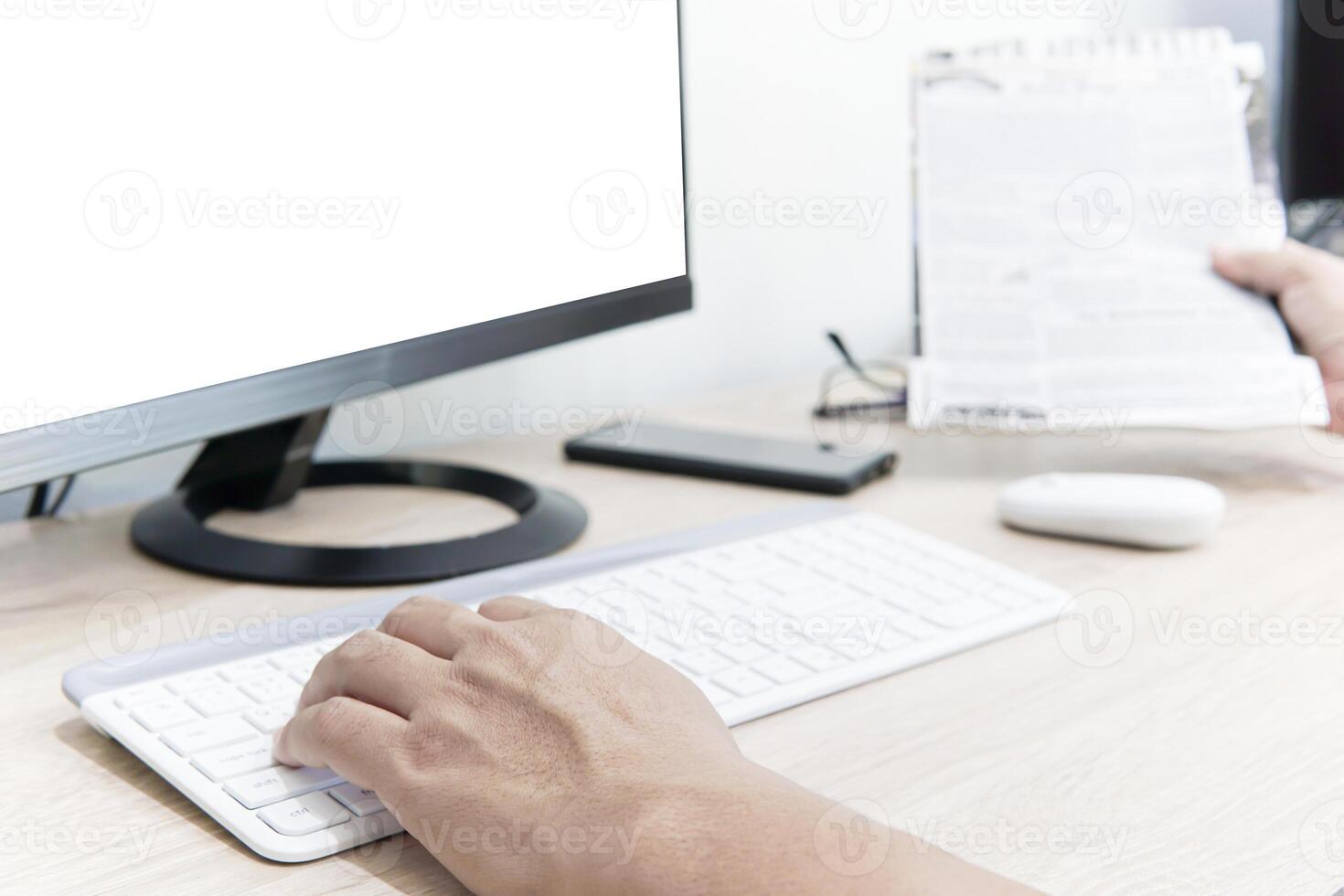 un hombre utilizar computadora escritorio buscar Noticias en el Internet con mirando un Noticias papel en el madera mesa foto