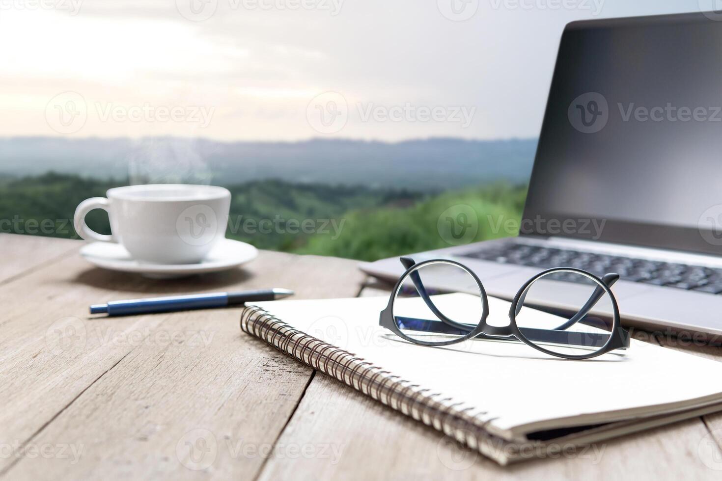 Glasses on paper notebook and coffee mug with laptop and wood table in mountain background photo
