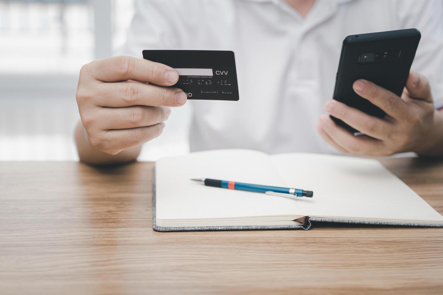 A man hold credit card and a smartphone in hand looking smart phone with shopping online and pencil notebook paper on wood table photo