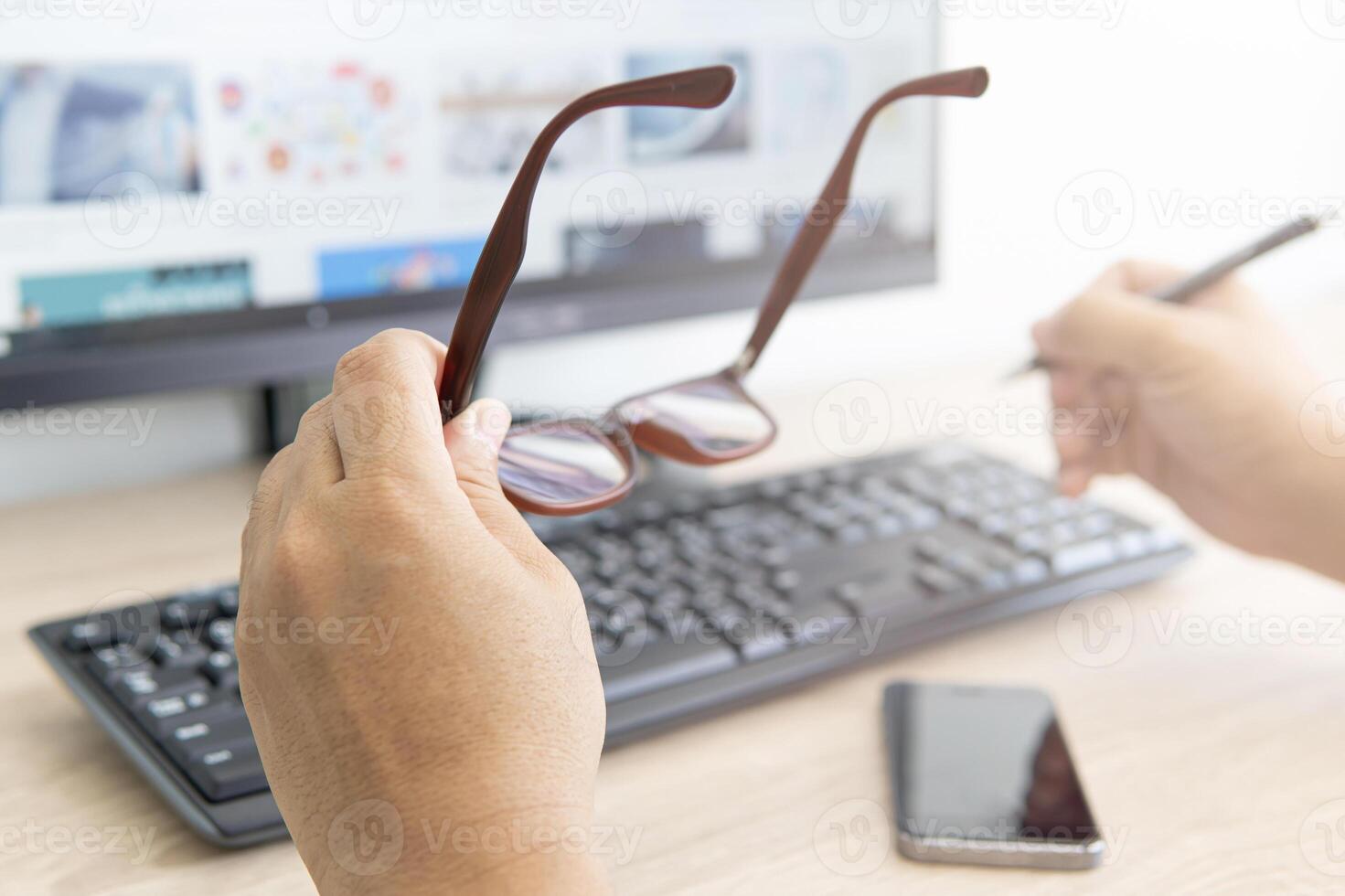 A man taking off glasses, feeling eye strain after long work with desktop computer and use a pen in the hand and smartphone on the wood table photo