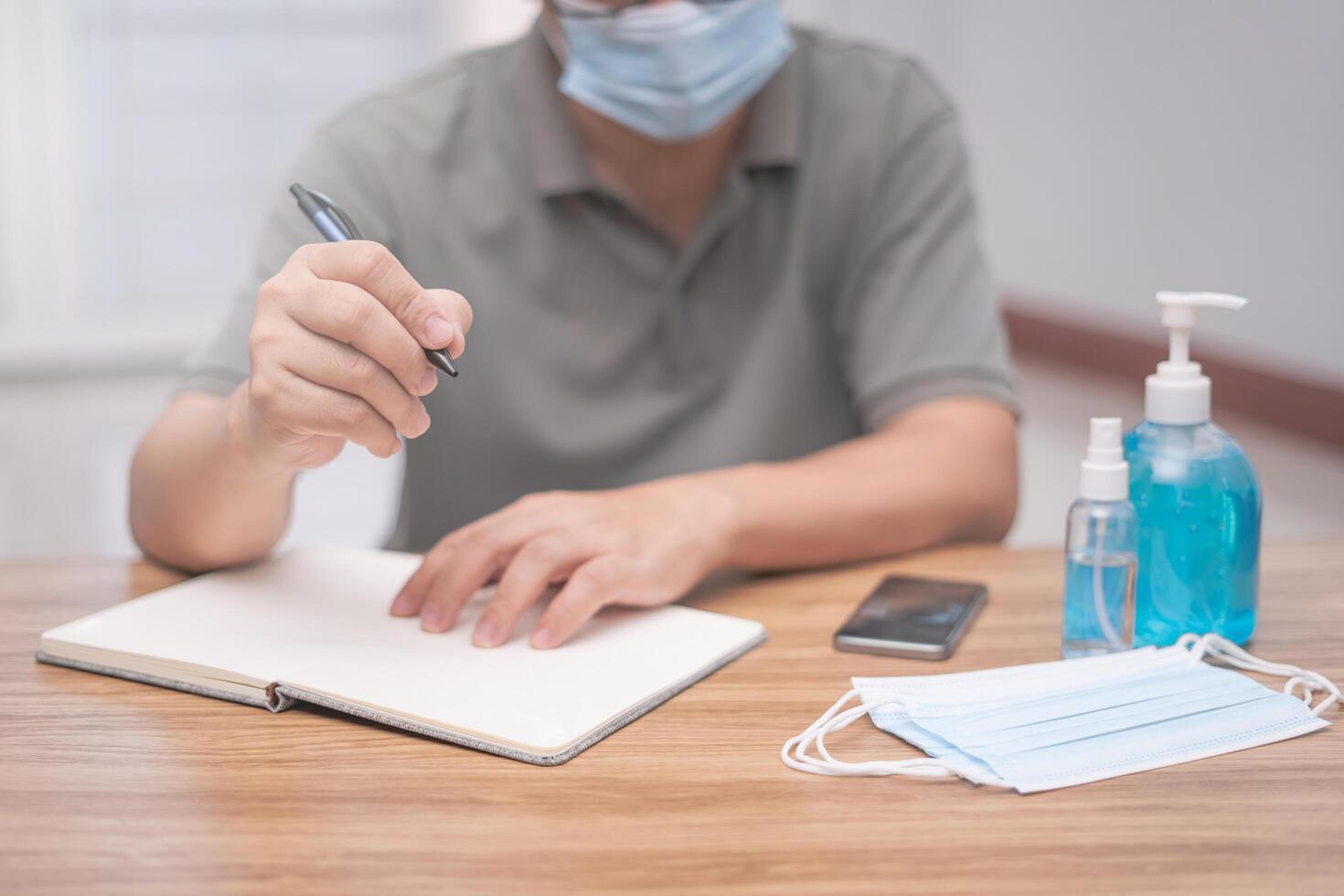 Work from home concept A man hold a pen write on the book and smartphone and Alcohol gel,Alcohol spray,Face mask on the wood table basic Antivirus protection concept photo