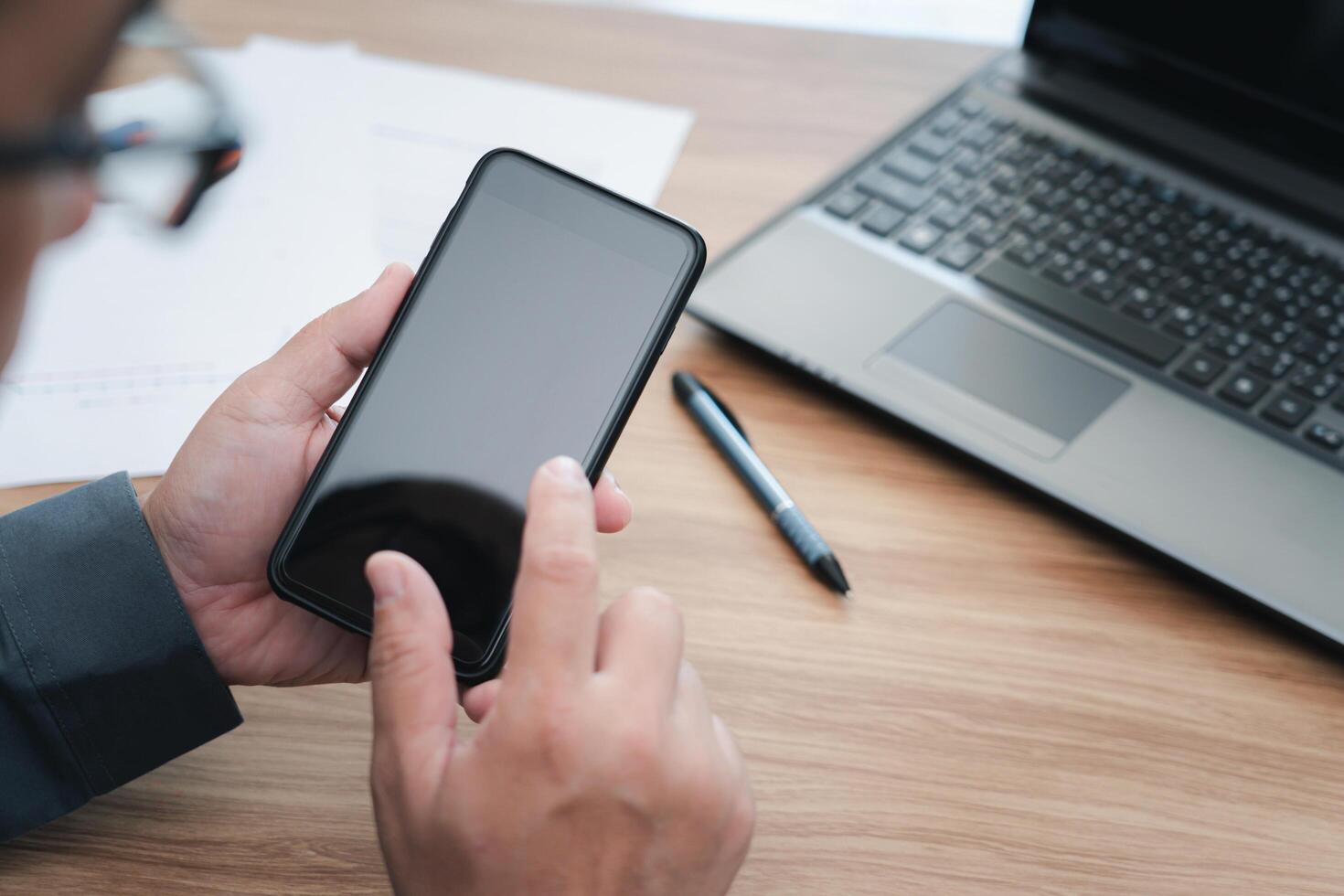 Close up display of smartphone in hand business man and laptop computer with wood table in background photo