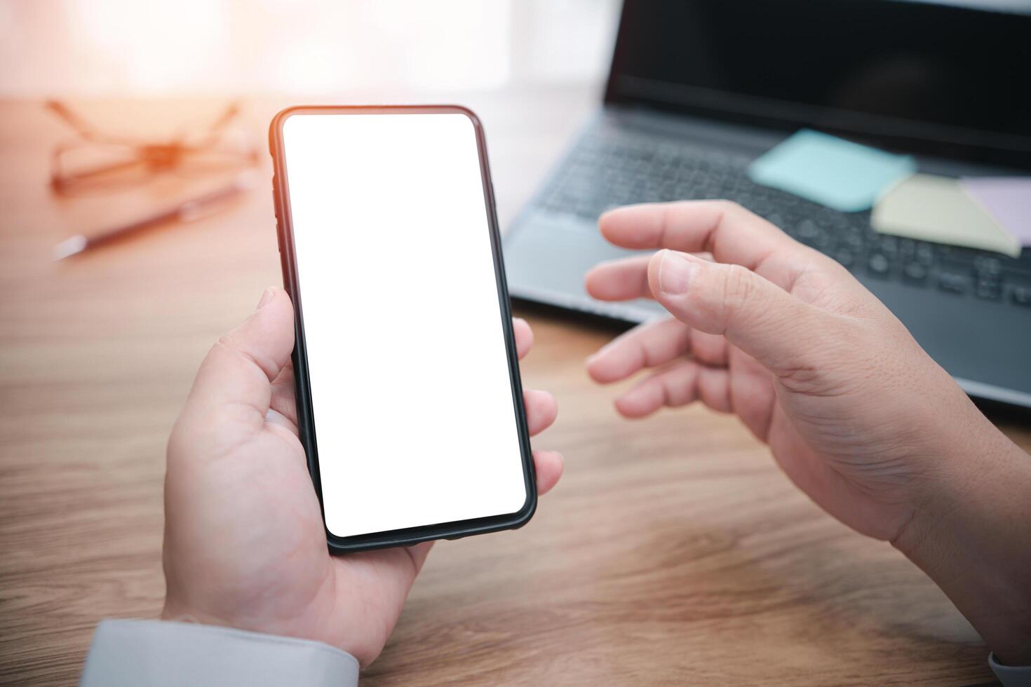 Close up white display of smartphone in hand business man and laptop computer with wood table in background photo