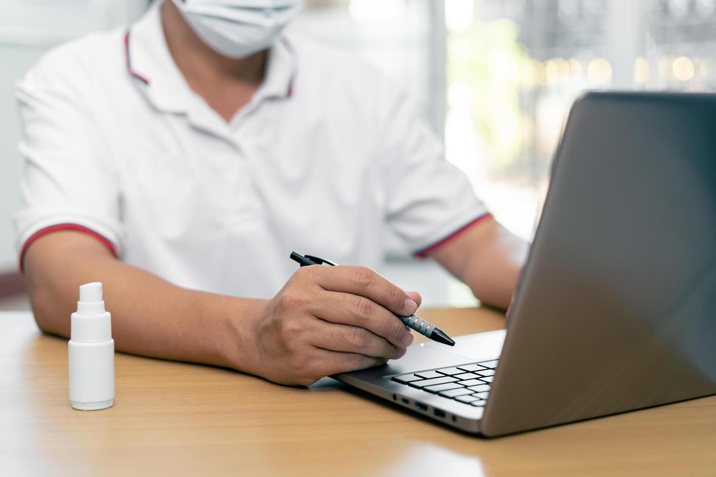 Work from home concept  Asia man wear a mask use laptop computer Find out about the spread of disease and use an clean and anti-virus alcohol spray on the wood table photo