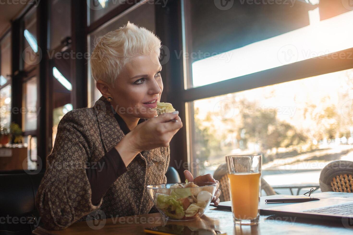 mayor mujer de 50 años es hermosa y elegante, se sienta en cafetería, trabajos con computadora portátil, Lanza libre diseñador. mira excelente, contento vida, activo estilo de vida. almuerzo, bocadillo, sano alimento, ensalada. foto