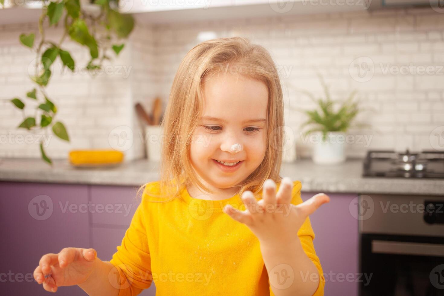 tres años niña en amarillo chaqueta es preparando a cocinar comida en cocina. retrato de contento niño. mano en harina. foto