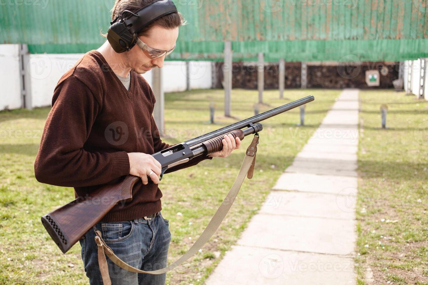 A man charges a pump-action shotgun with a Ammo. 12 caliber. Tyre outdoor. A man in headphones and goggles is preparing to shoot. Firearms for sports shooting, hobby. photo