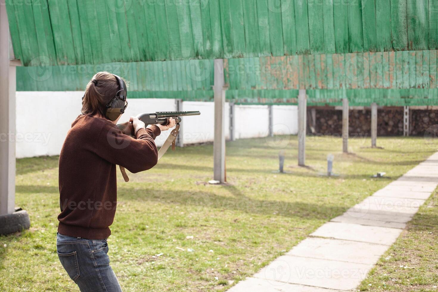 A man shoots at metal flags, targets. Firearms pump-action shotgun. Tyre's outdoor. Patrons with a fraction. Green grass, a man with headphones, a glade with protection for shooting. copy space photo