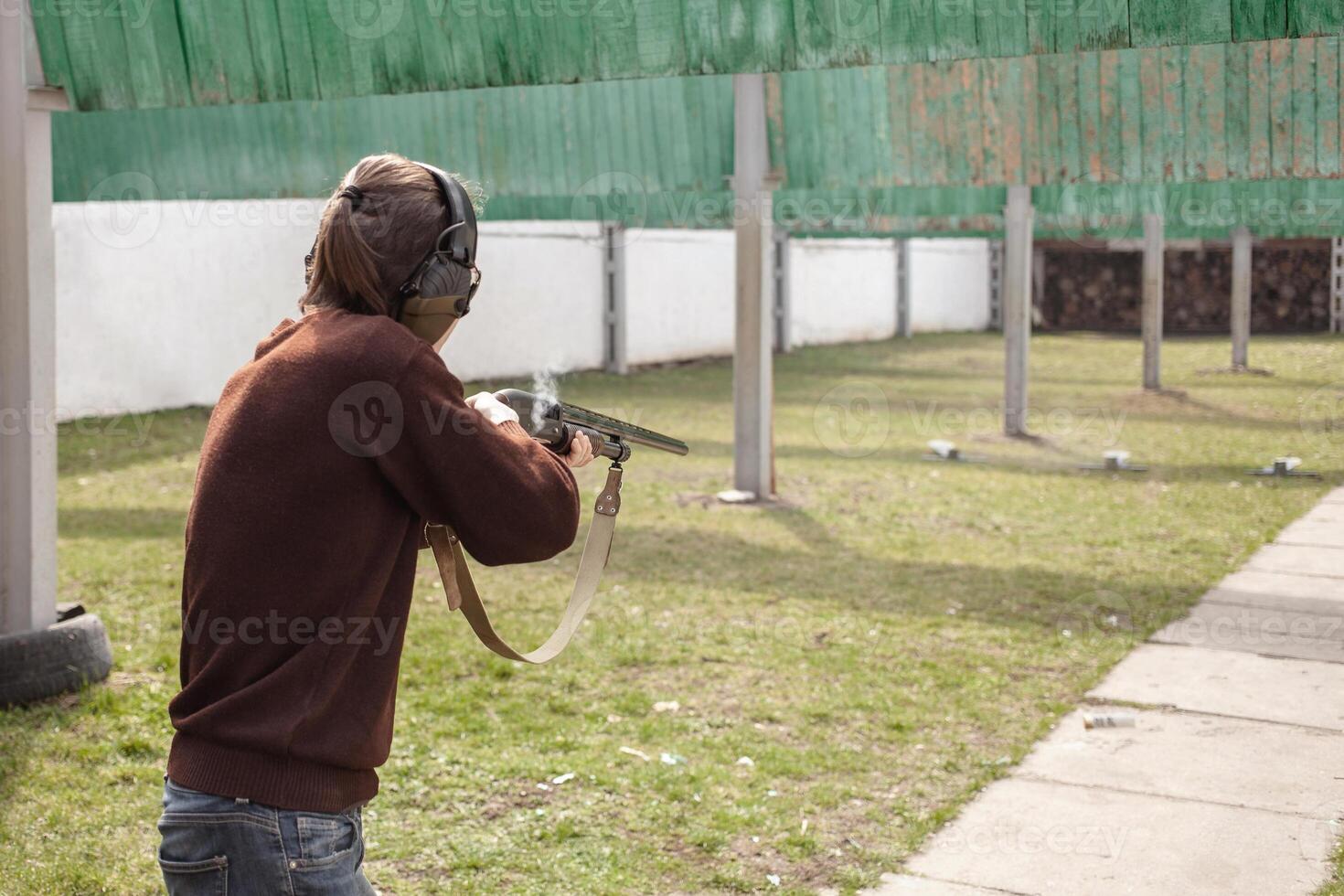 A man shoots at metal flags, targets. Firearms pump-action shotgun. Tyre's outdoor. Patrons with a fraction. Green grass, a man with headphones, a glade with protection for shooting. photo