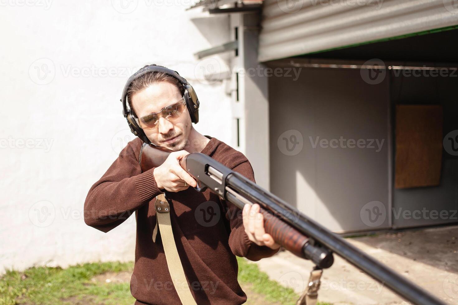 A man with a pompous firearm on a white background. A man in protective glasses and headphones. Tyre outdoor. Grey hangar. gun photo