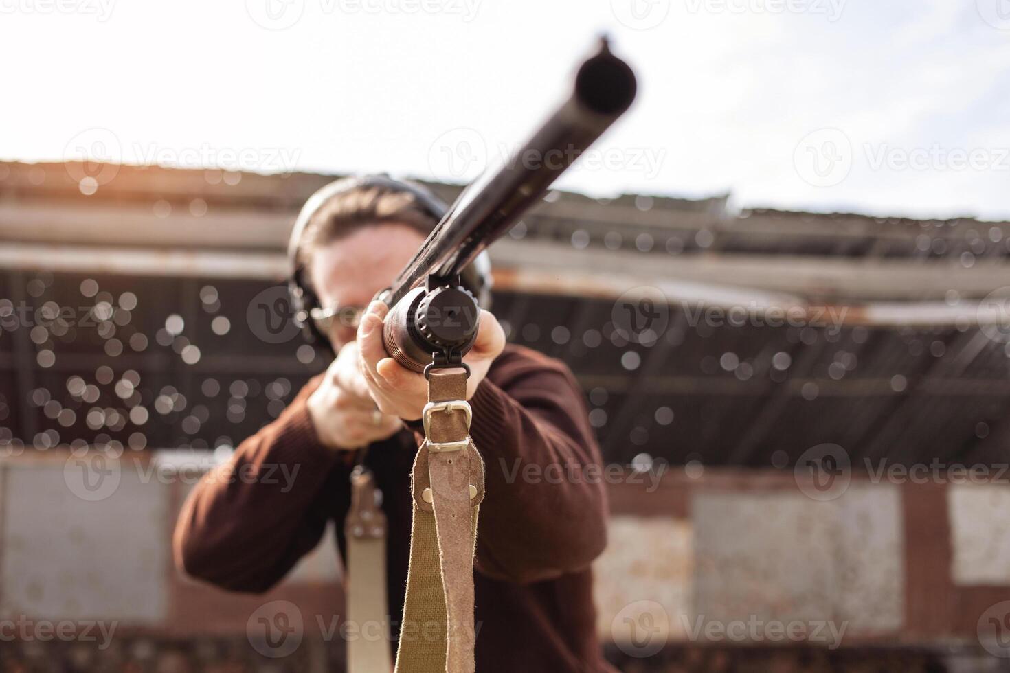 A man in protective glasses and headphones. A pump-action shotgun. Tyre's outdoors. A wall and a roof with bullet holes. Sport photo