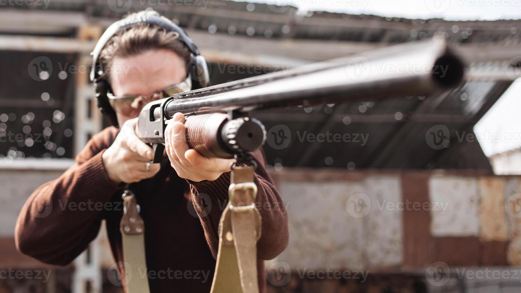 A man in protective glasses and headphones. A pump-action shotgun. Tyre's outdoors. A wall and a roof with bullet holes. Sport photo