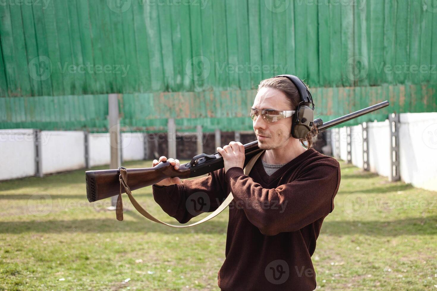 A man in protective glasses and headphones. A pump-action firearm shotgun. Tyre's outdoors. Green grass, protection for sports shooting. hobby. close up photo