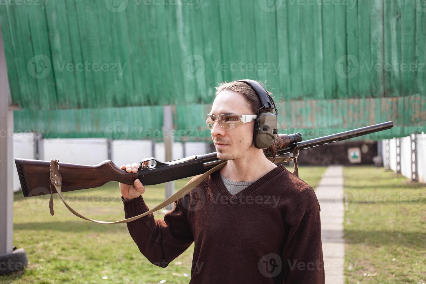 A man in protective glasses and headphones. A pump-action firearm shotgun. Tyre's outdoors. Green grass, protection for sports shooting. hobby. photo