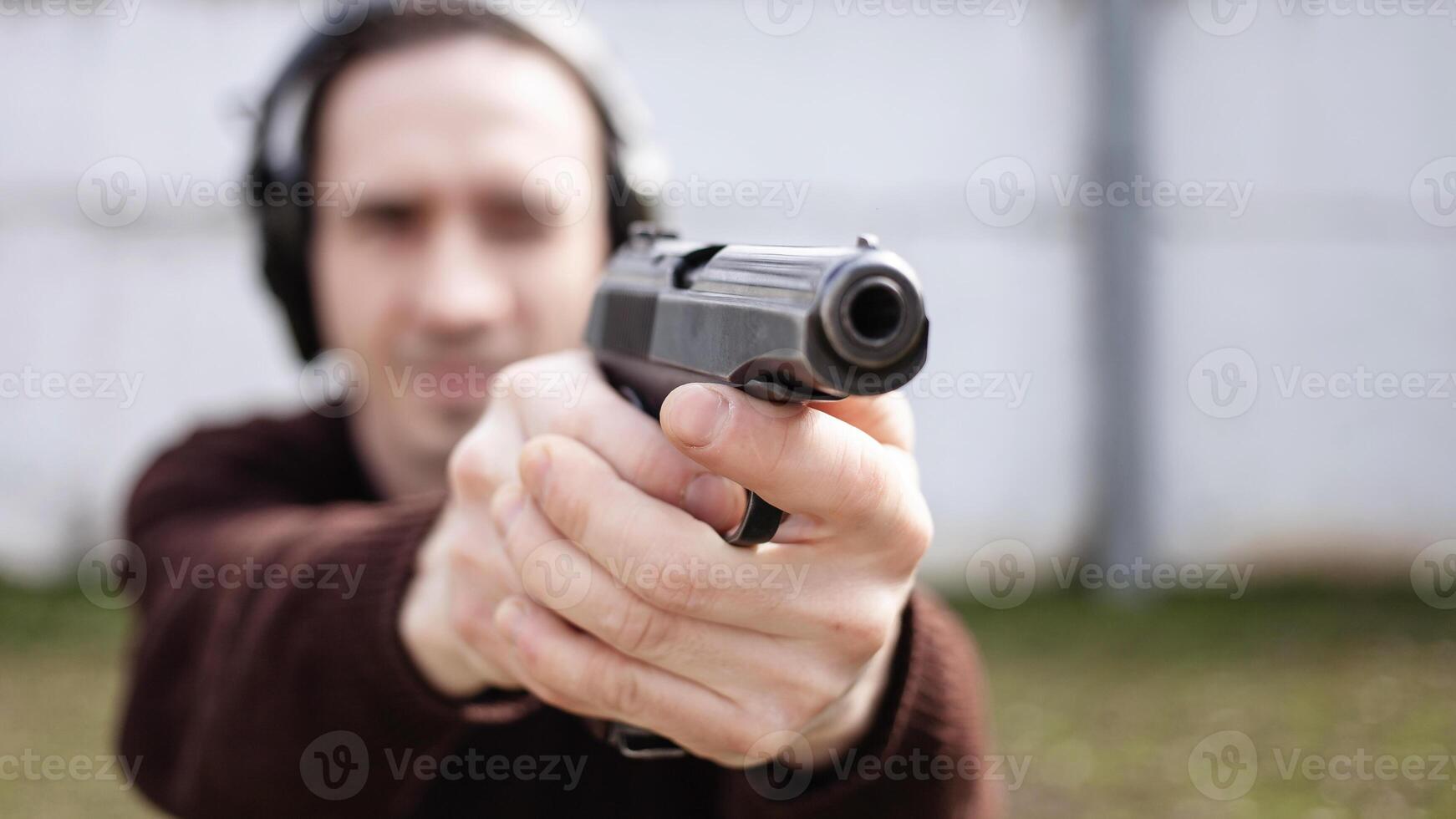 A man is aiming for a gun. A man wearing protective headphones. outdoor tyre shooting range. hobby. firearm against the white wall photo