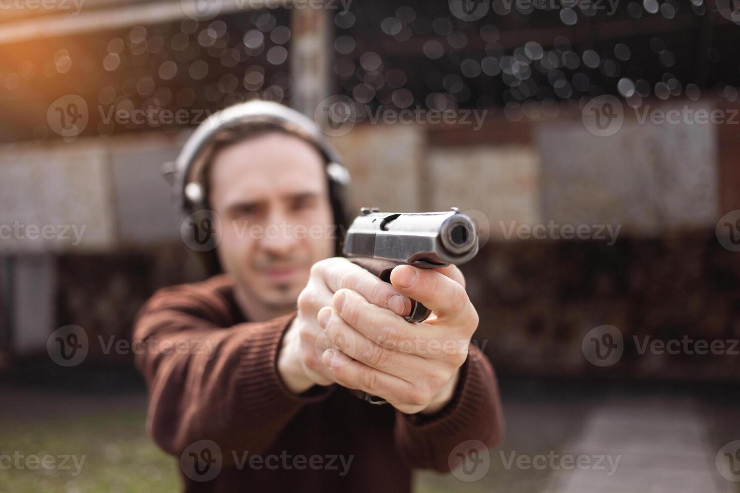 A man shoots a gun, aiming at the target. A man wearing protective headphones. A wall and a roof with bullet holes. outdoor shooting range photo