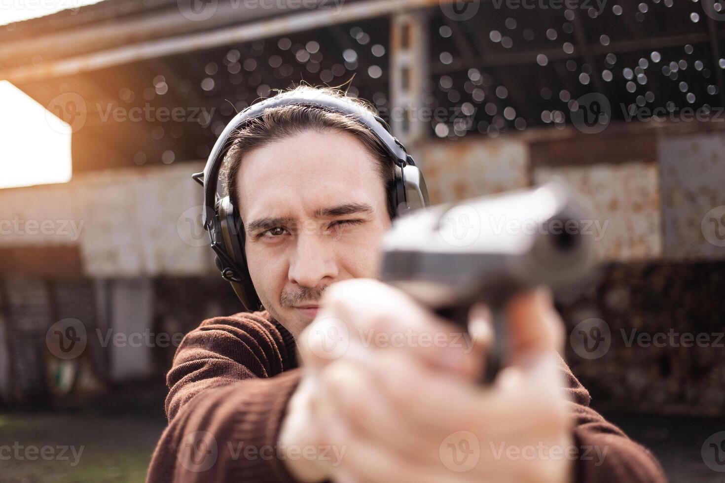 A man shoots a gun, aiming at the target. A man wearing protective headphones. A wall and a roof with bullet holes. outdoor shooting range close up photo