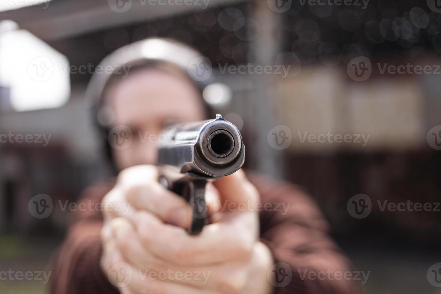 A man shoots a gun, aiming at the target. A man wearing protective headphones. A wall and a roof with bullet holes. outdoor shooting range photo