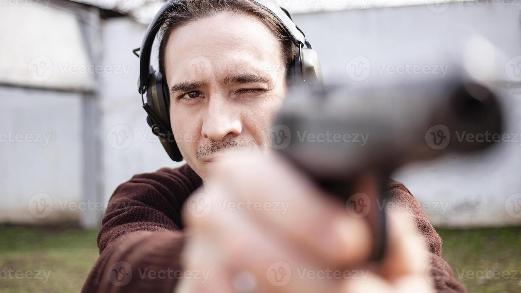 A man is aiming for a gun. A man wearing protective headphones. outdoor tyre shooting range. hobby. firearm against the white wall close up photo